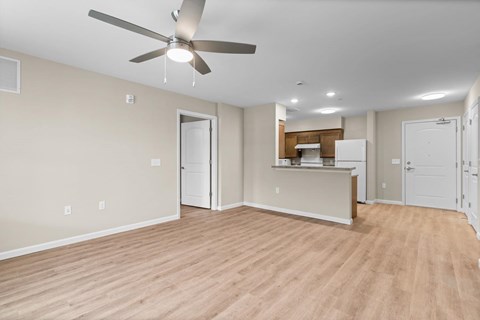 A room with a ceiling fan and light fixture, wooden flooring, and a kitchenette in the background.
