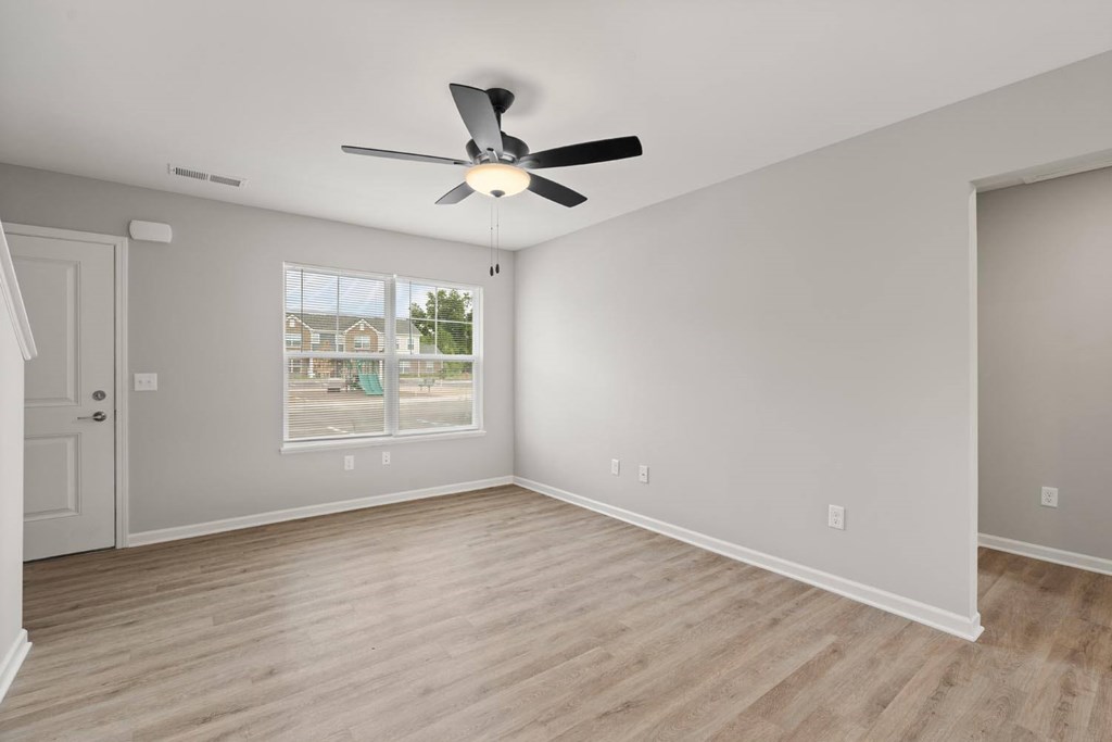 A room with a ceiling fan and light fixture, wooden flooring, and a window with a view of a street and buildings outside.