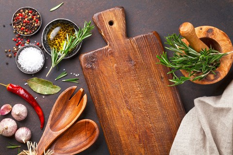 A wooden cutting board with a handle is surrounded by various herbs and spices.