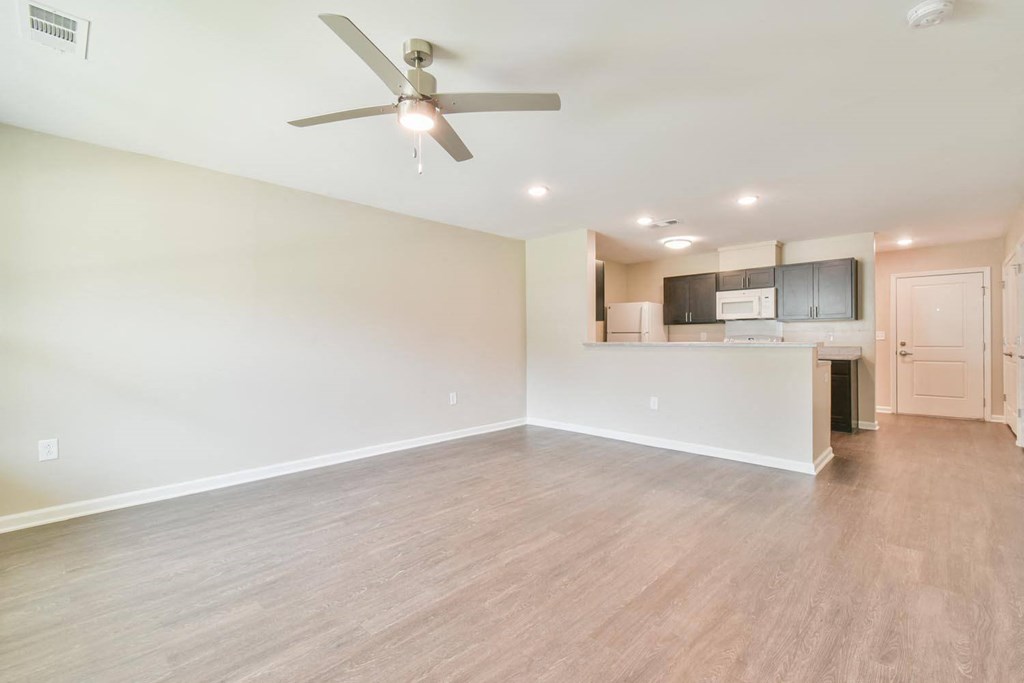 an empty living room with a ceiling fan and a kitchen in the background