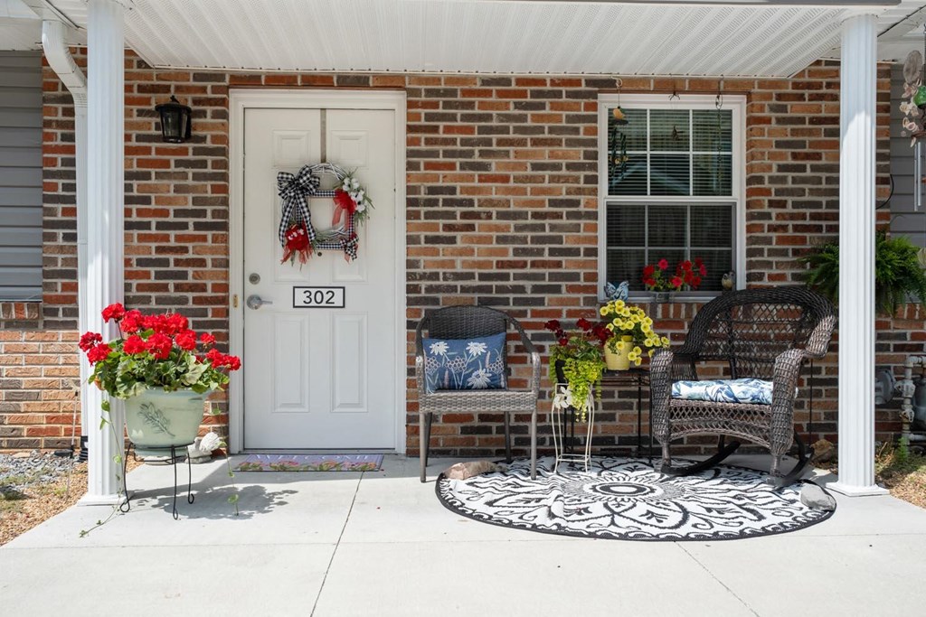 the front porch of a brick house with two wicker chairs and a white door