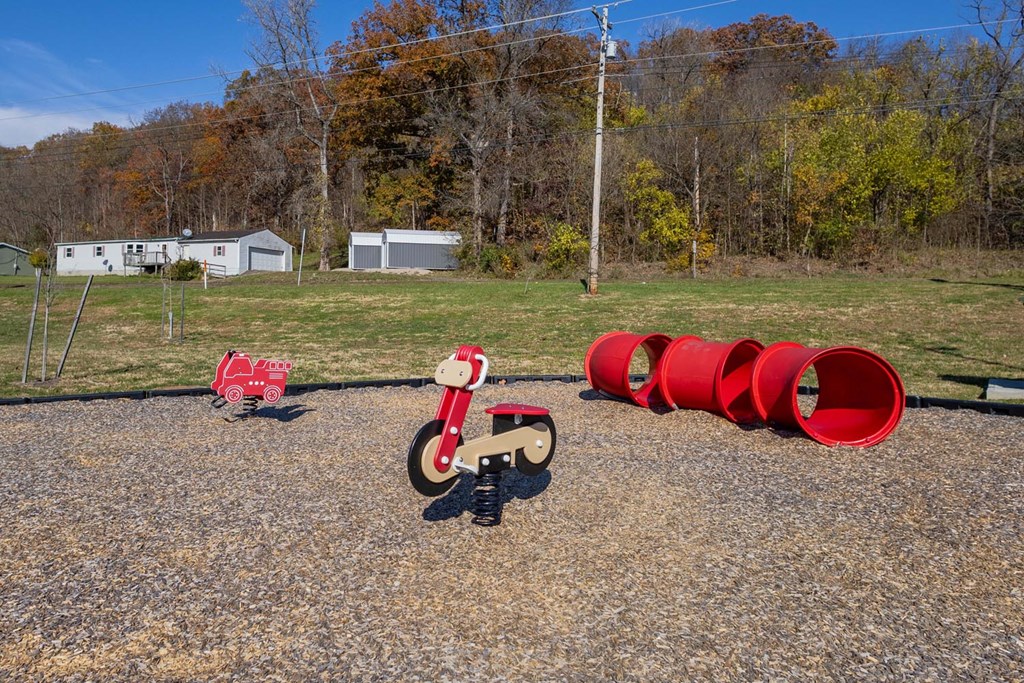 A red toy motorcycle is in the foreground of a gravel area with a few red barrels and a few buildings in the background.