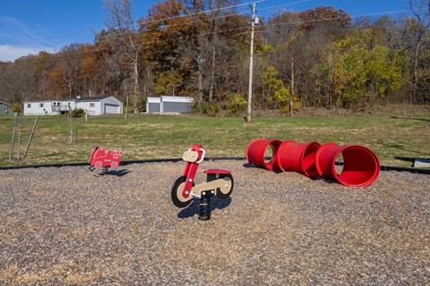 A red toy motorcycle is in the foreground of a gravel area with a few red barrels and a few buildings in the background.