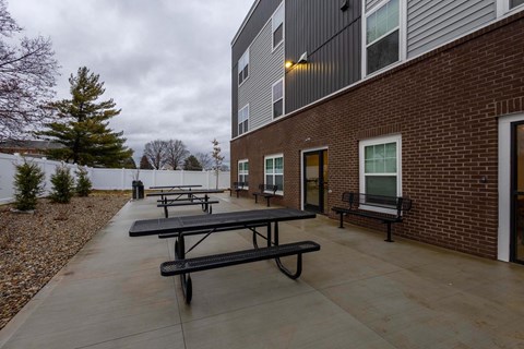 A series of black picnic tables are lined up on a concrete patio outside of a brick building.