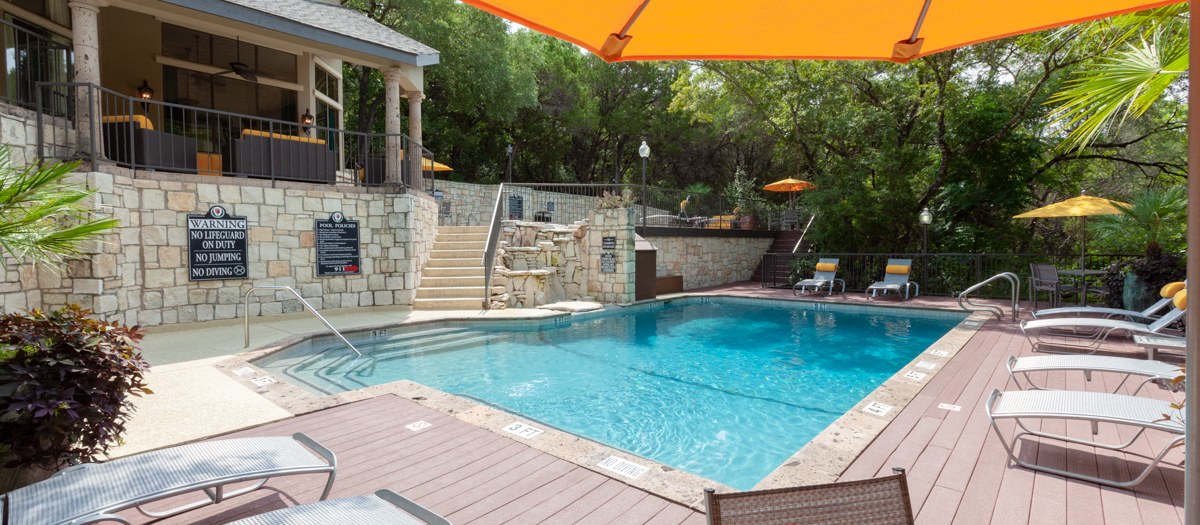 A pool with a stone wall and a patio with chairs and umbrellas.