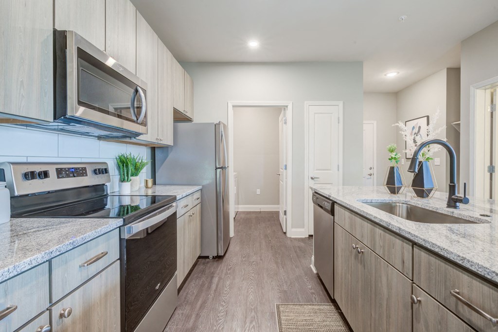 one bedroom kitchen with gooseneck black faucet on the island,  woodgrain light cabinets, stainless steel appliances, granite countertops, kitchen table with blue leather chairs, 6-panel white doors, woodlike plank flooring, and laundry room off the kitchen