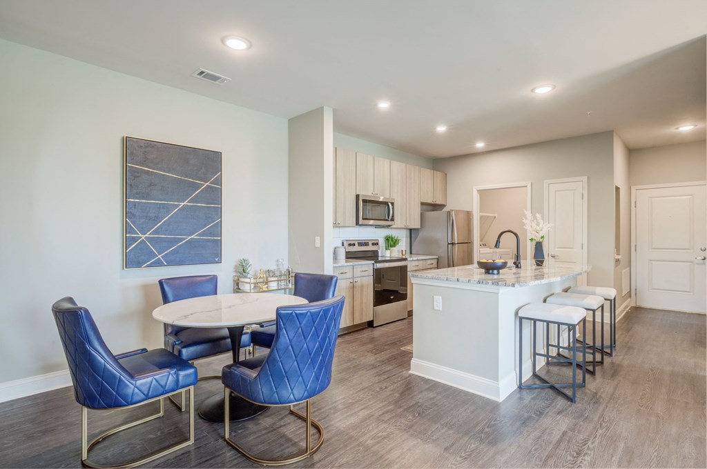 one bedroom kitchen with woodgrain light cabinets, stainless steel appliances, granite countertops, kitchen table with blue leather chairs, 6-panel white doors, woodlike plank flooring, and laundry room off the kitchen