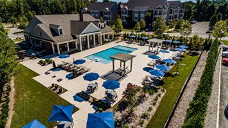 Over look of pool and club house. Multiple patio seating areas with umbrellas, pool, club housebuilding, parking lot, trees, and pavilion