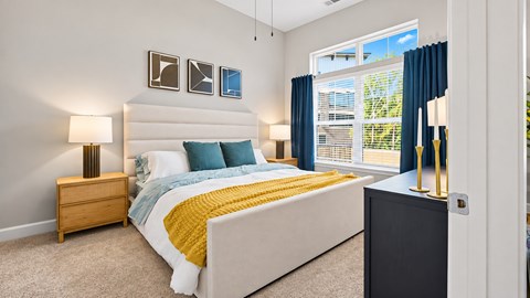 Furnished apartment bedroom with an upholstered bed, blue curtains and a large window at Argento at Sugarloaf.