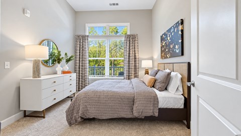 Furnished apartment bedroom with gray bedding, a white dresser and patterned curtains at Argento at Sugarloaf.