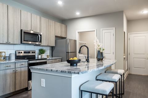 Model kitchen with granite countertops featuring a grey and white marble effect. there are stainless steel appliances including microwave, stove and fridge. The gooseneck sink faucet is black.  the cabinets are light woodgrain and the flooring is a wood-like plank.