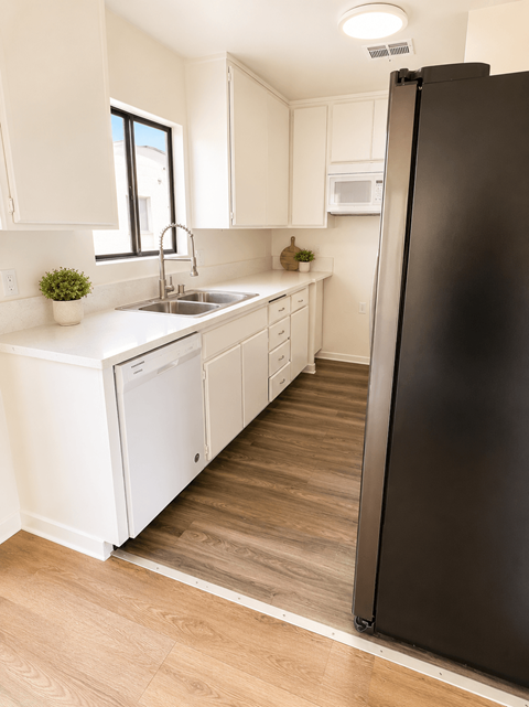 A black refrigerator stands in a kitchen with white cabinets and a wooden floor.