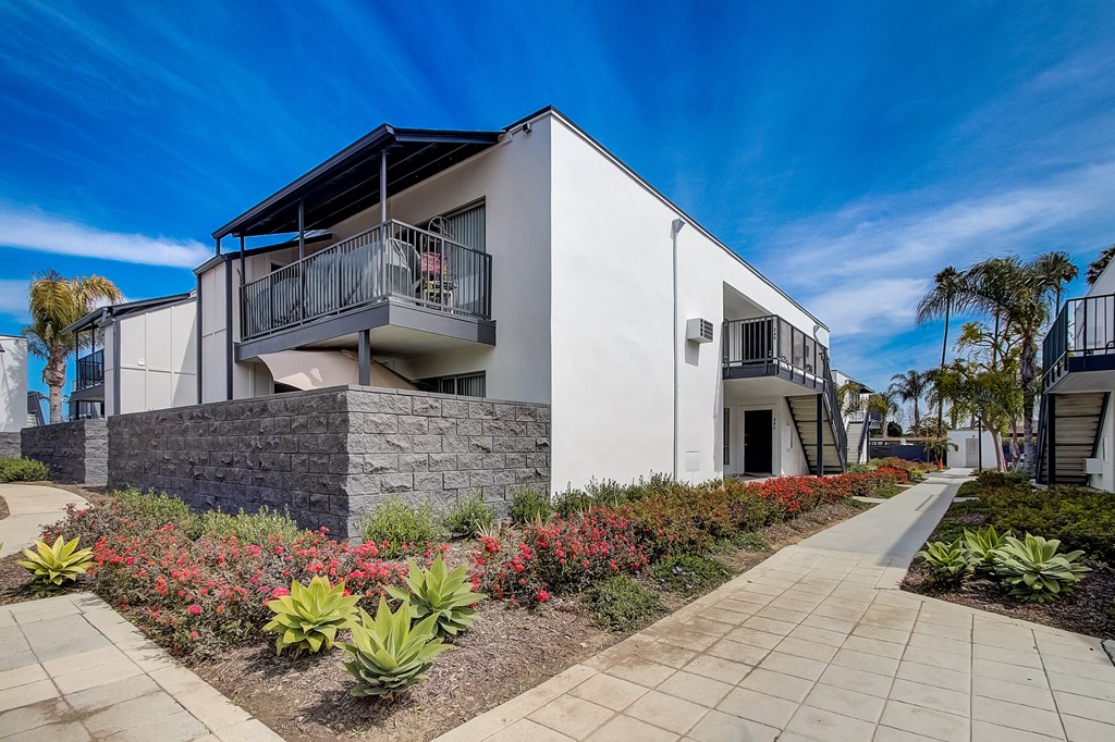 a house with a white facade and a stone wall with flowers and bushes in front of it