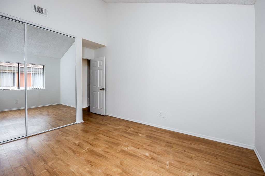 the living room and bedroom of an apartment with wood flooring and a mirrored closet