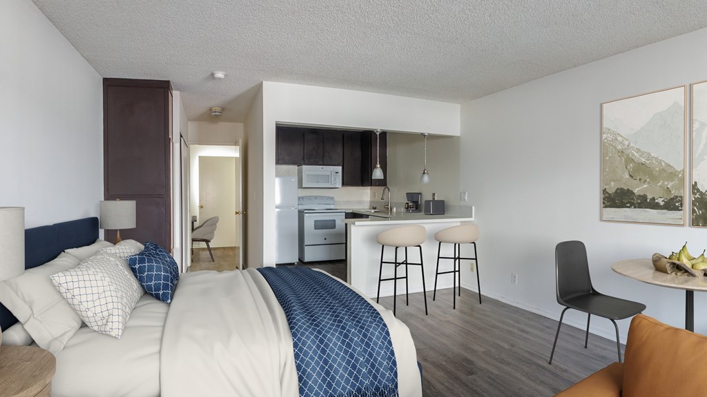A bedroom with a bed, chair, and a kitchen area in the background.
