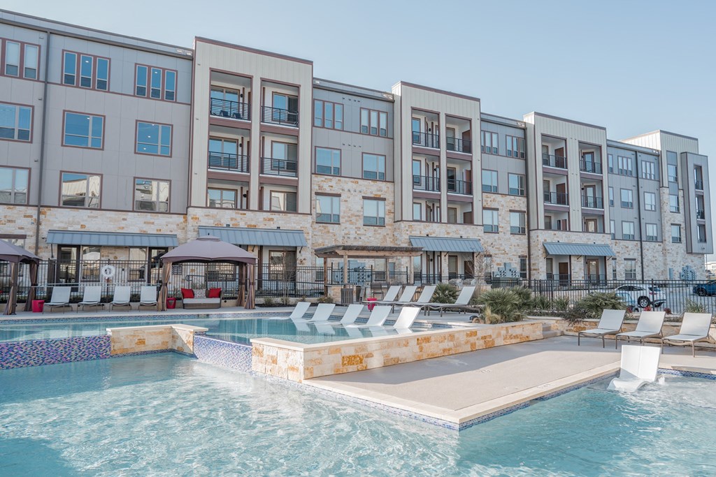 Apartments College Station TX - Caprock Crossing - Outdoor Expansive Pool, with Stone Details, Different Levels, and Submerged Seating, Surrounded by Lounge Chairs, Covered Cabanas with Benches, and Manicured Plant Beds
