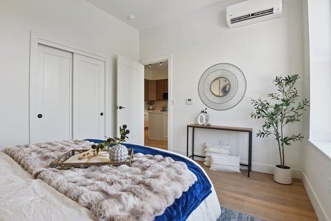 A bedroom with a bed covered in a grey fur throw, a round mirror on the wall, and a small potted plant on the floor.
