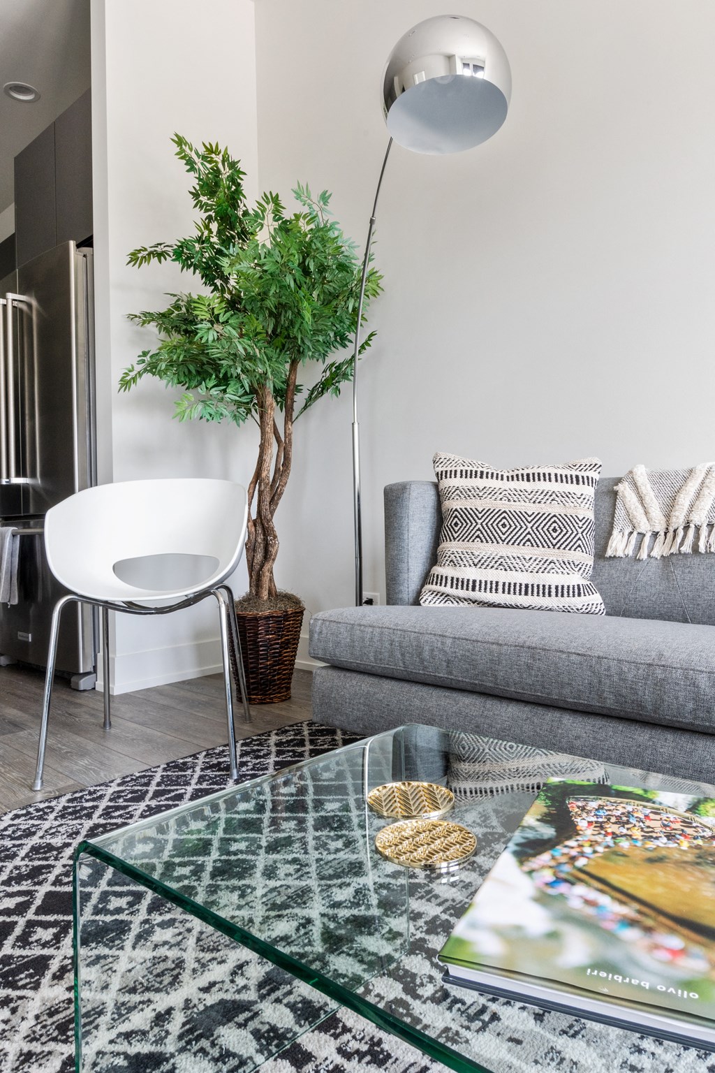 A part of the living room with a gray couch, glass coffee table, floor lamp, and huge plant in the corner
