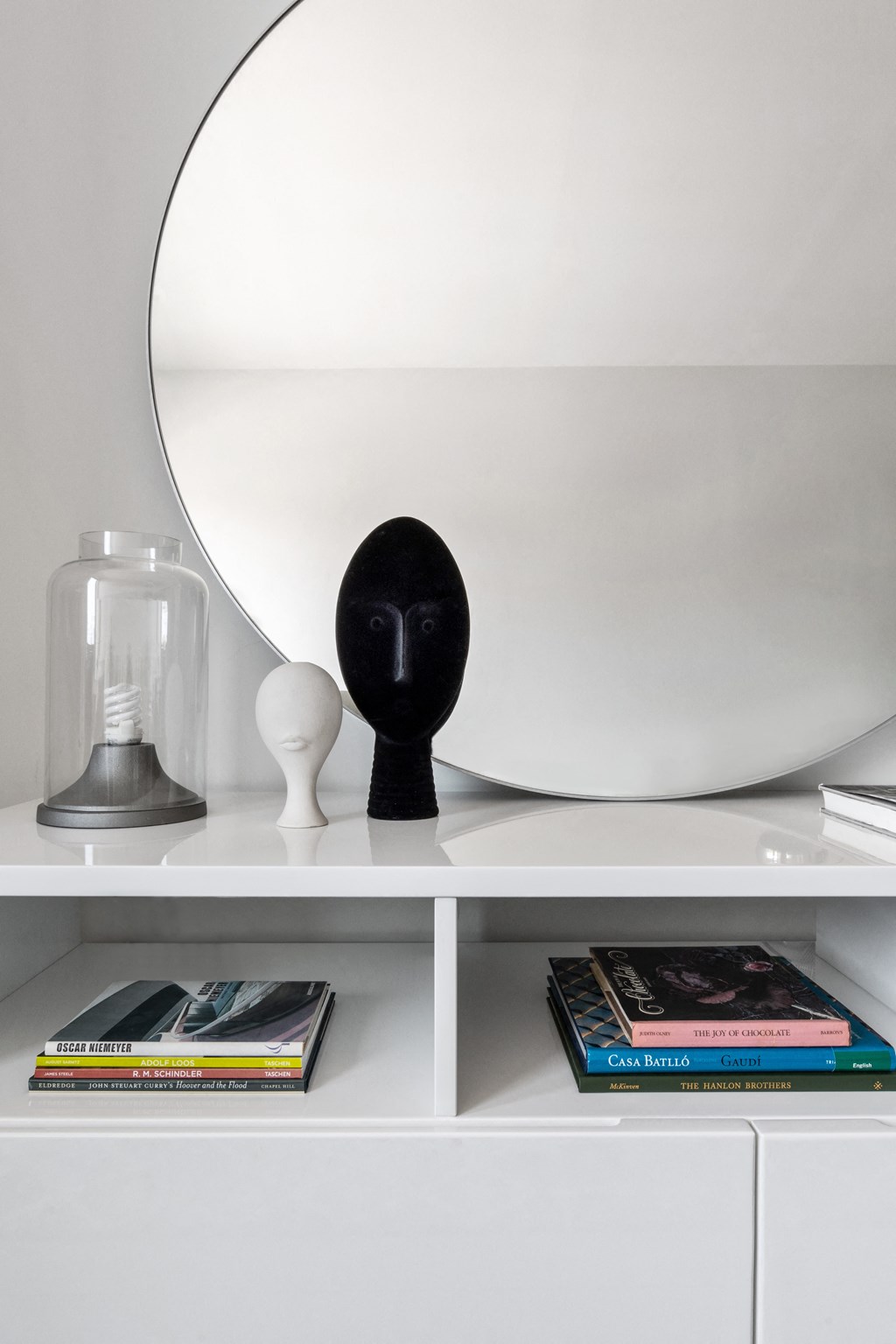 A round mirror on a white shelf with books and sculptures