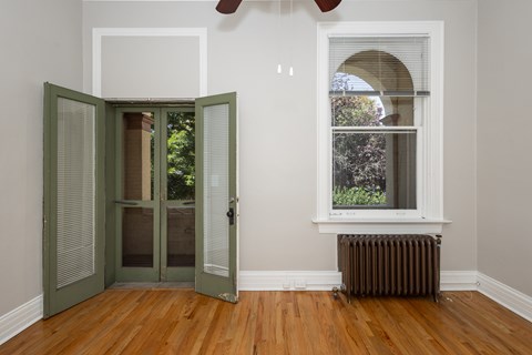 A room with a wooden floor, a window with a view of trees, and a wall with a white frame.