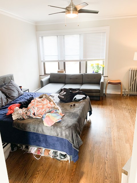 A messy bedroom with a bed covered in clothes and a couch in the background.