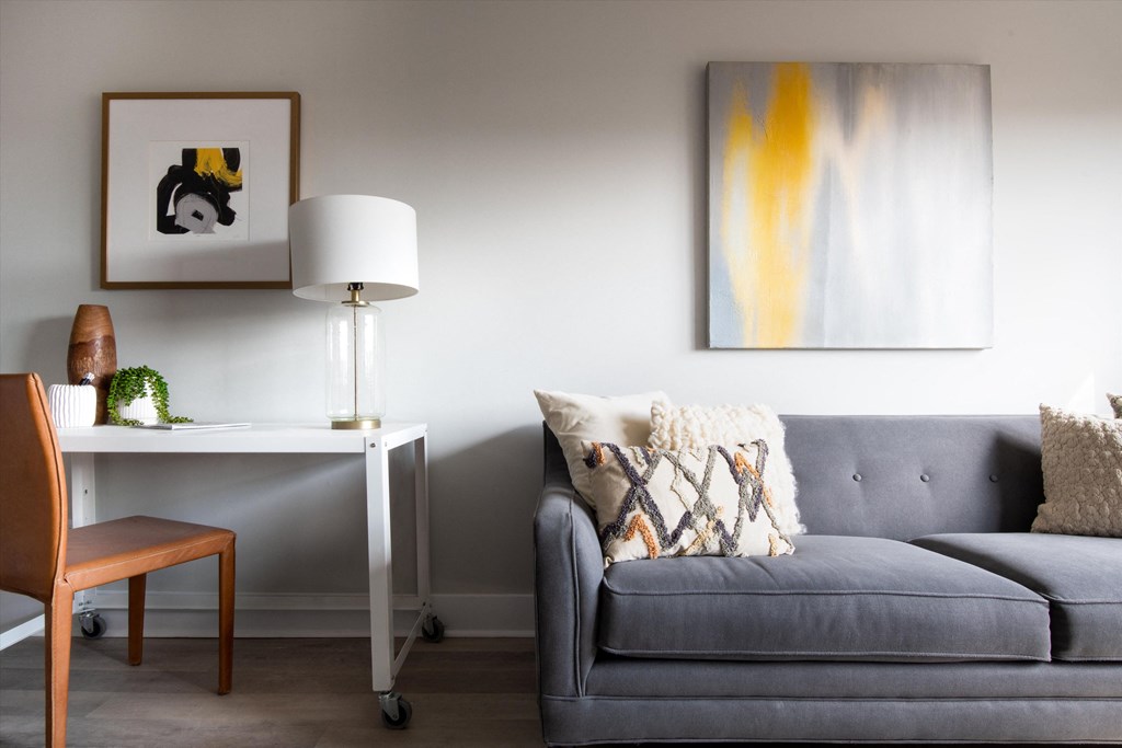 a living room with a grey sofa and a white desk with a lamp and a painting on