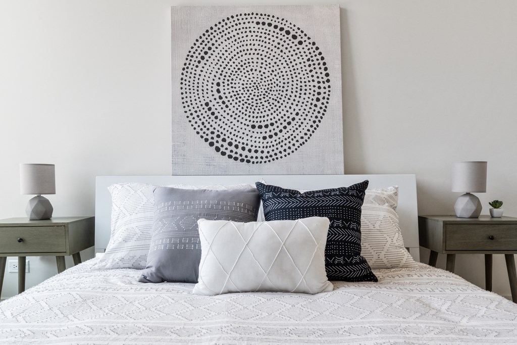 A bedroom with white bedding, cozy pillows, and an abstract painting on the wall