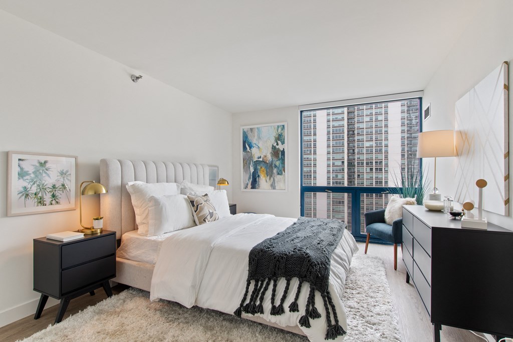 A bedroom with a king size bed, two night stands, and large floor to ceiling window in the background