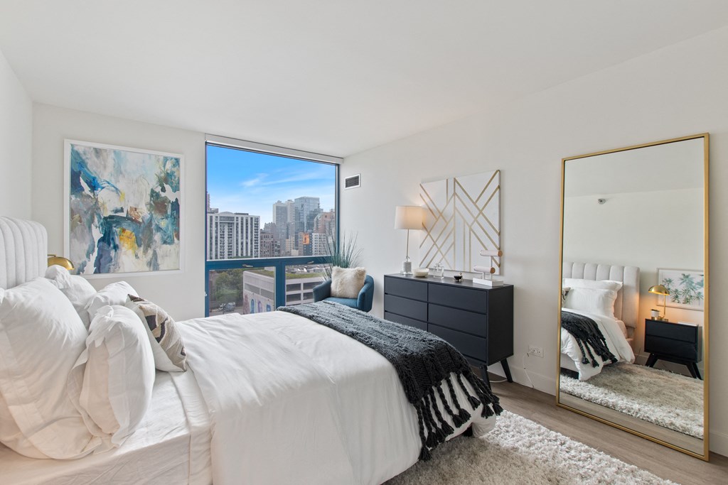 A bedroom with a large bed, gray modern dresser, large mirror, and a large window with the urban view