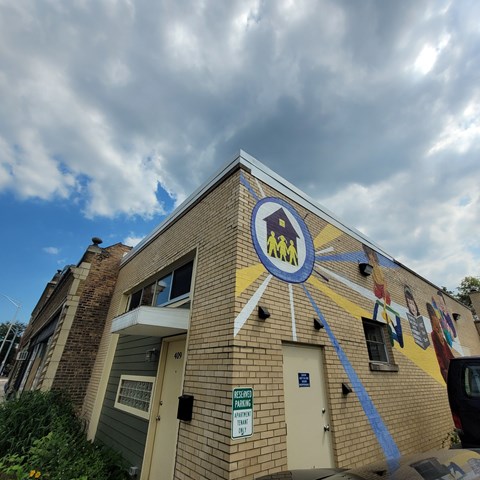 A building with a mural on the side has a blue sign on the front door.