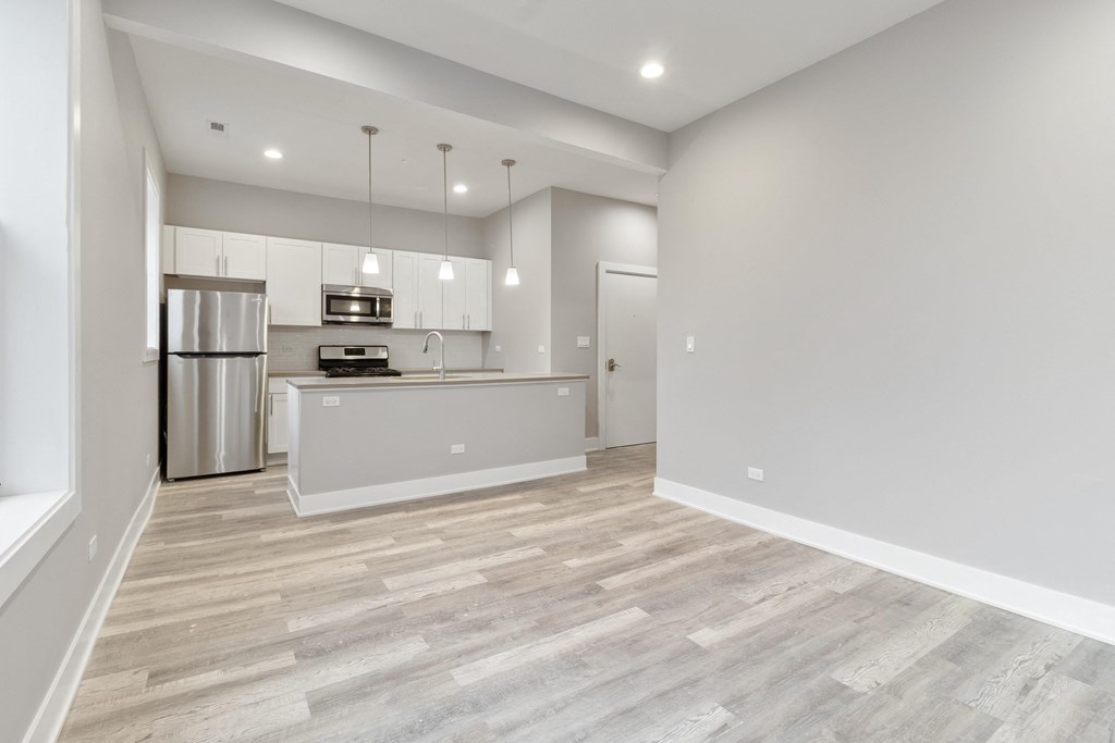 an empty living room with a kitchen with a stainless steel refrigerator