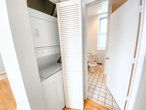 A white washing machine is in a small room with a toilet and a tiled floor.