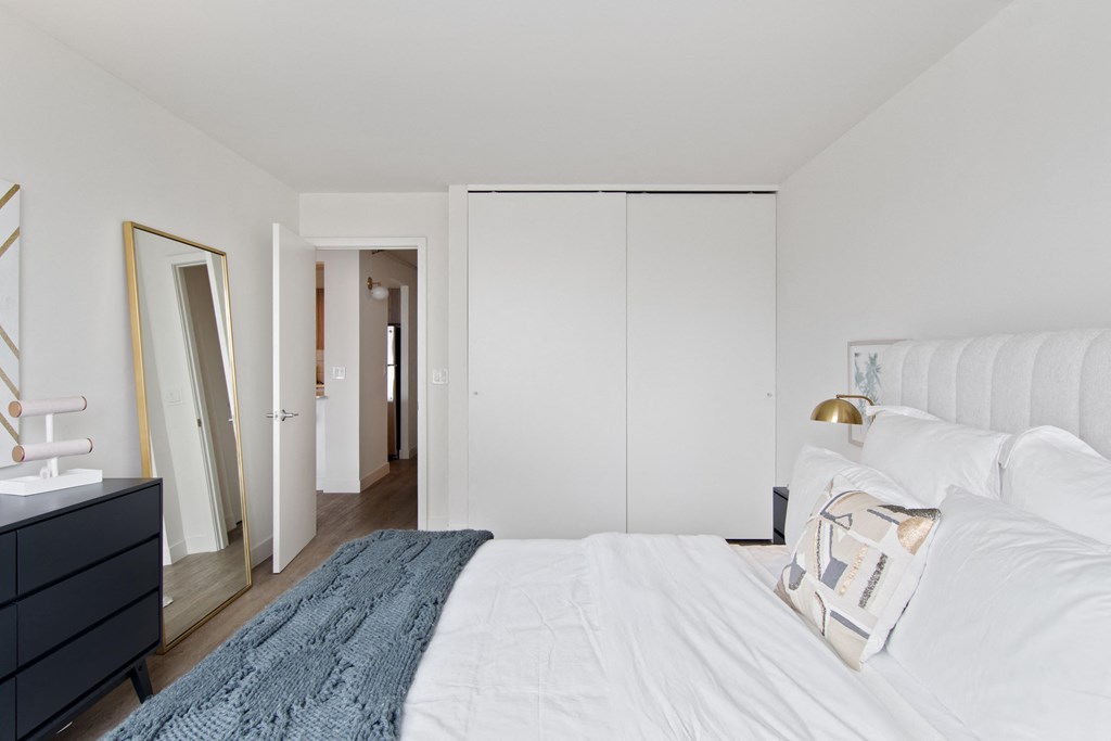 A bedroom with white walls and a bed with a blue blanket and white bedding, with a large closet in the backround
