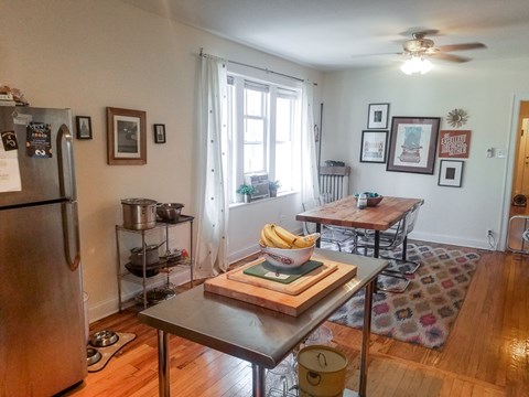 A kitchen with a refrigerator, a table with a bowl of bananas, and a rug on the floor.