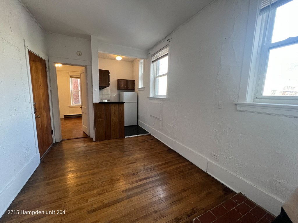 A room with wooden floors and white walls, with a kitchen area visible in the background.