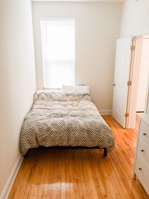 A bed with a white and black patterned cover sits in a room with wooden floors and white walls.