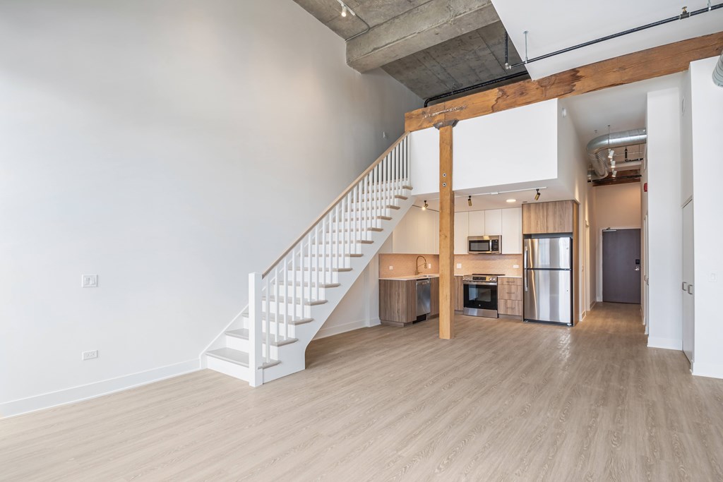 A white staircase with a wooden handrail leads to a living room with a kitchen in the background.