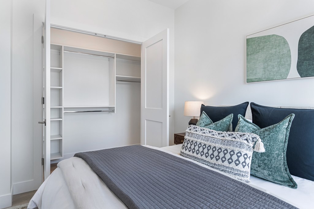 A bed with a grey blanket and a white pillow is in the foreground of a room with a white door and a lamp.