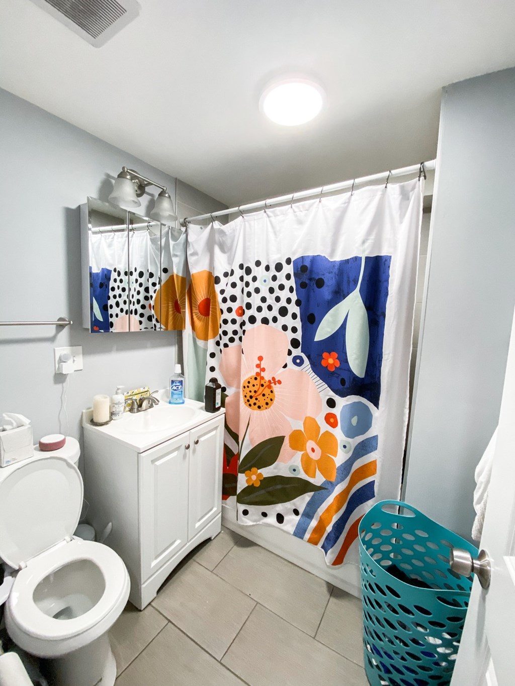 A bathroom with a toilet, a shower curtain with a floral pattern, and a green waste basket.