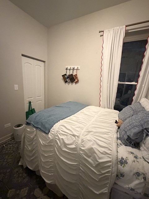 A bed with a blue blanket and a white pillow is in a room with a white door and a window with white curtains.