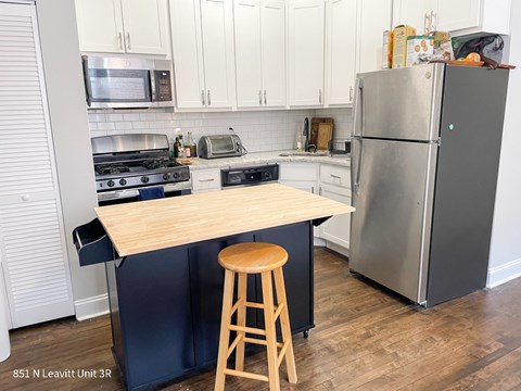 A kitchen with a stove top oven, microwave, toaster, refrigerator, and a wooden island with a stool.