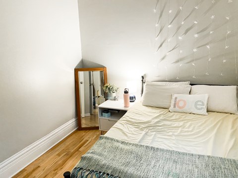 A white bed with a grey blanket and a white pillow with a red design on it.