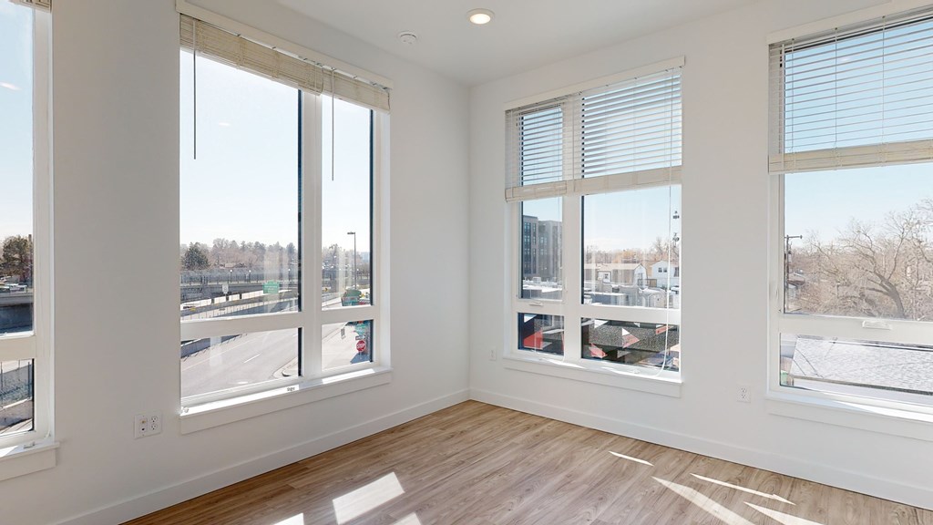 A room with white walls and wooden flooring, featuring a large window with a view of a parking lot and trees.