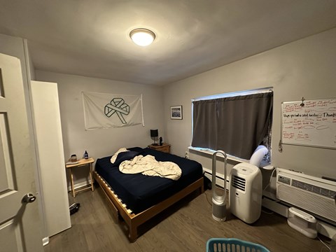 A bedroom with a bed, a fan, a window with curtains, and a whiteboard on the wall.
