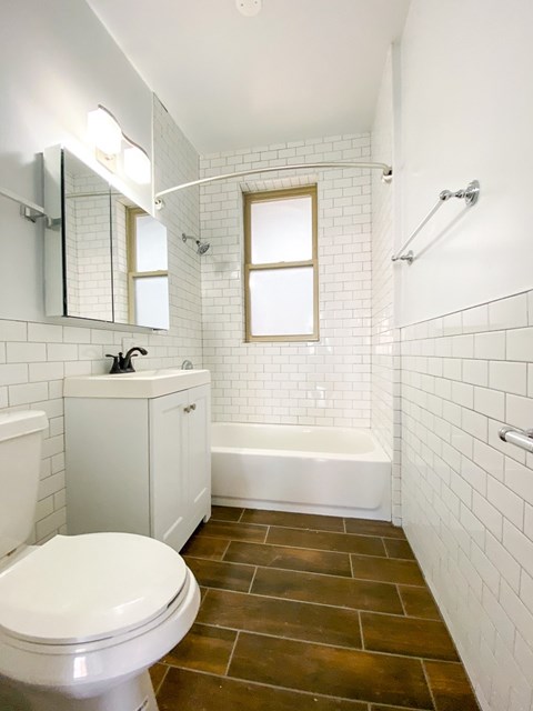 A white toilet sits in a bathroom with a white tub and a window.