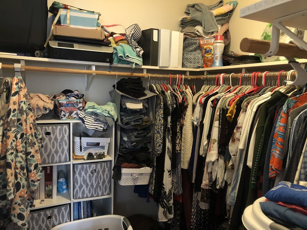 A cluttered closet with clothes and a computer monitor on the shelf.