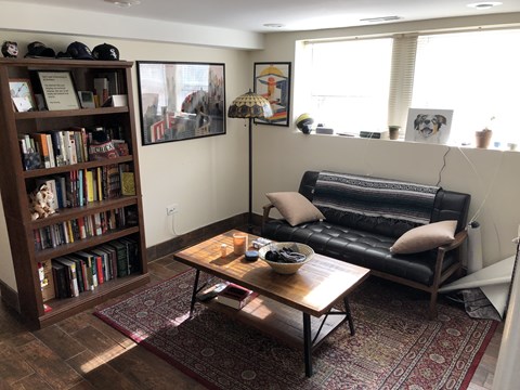 A living room with a black leather couch, a wooden bookshelf filled with books, a coffee table with a bowl of fruit on it, and a rug on the floor.