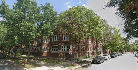 A street view of a residential area with a red brick building on the left and cars parked on the right.