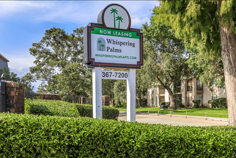 A sign for Whispering Palms apartments is displayed in front of a building.