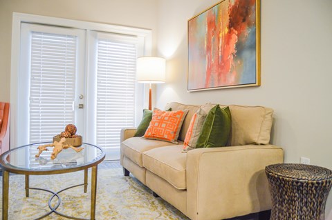 A living room with a beige couch, a glass table, a lamp, and a painting on the wall.